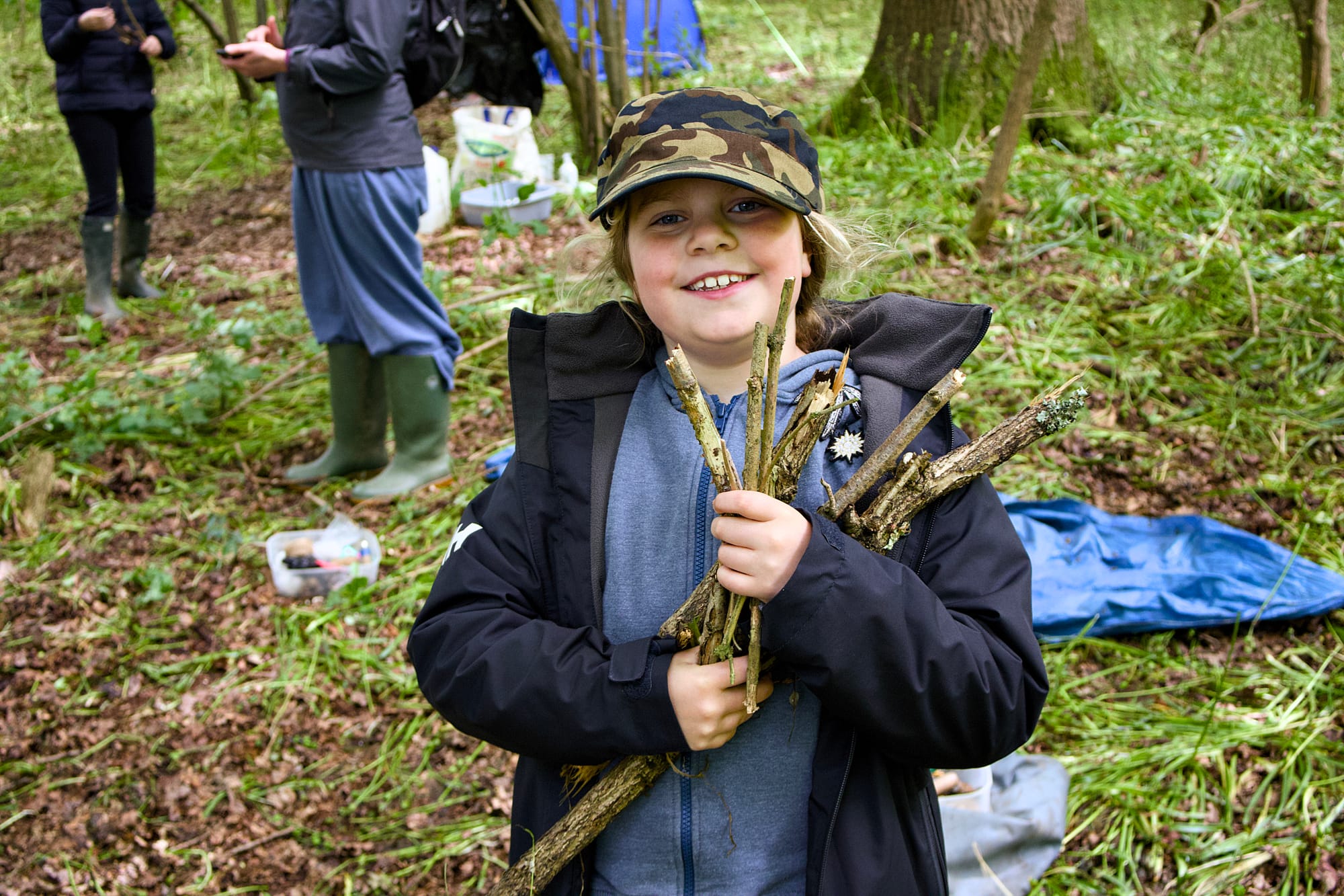A nine-year old girl in outdoors clothes, clutching sticks in a forest school setting